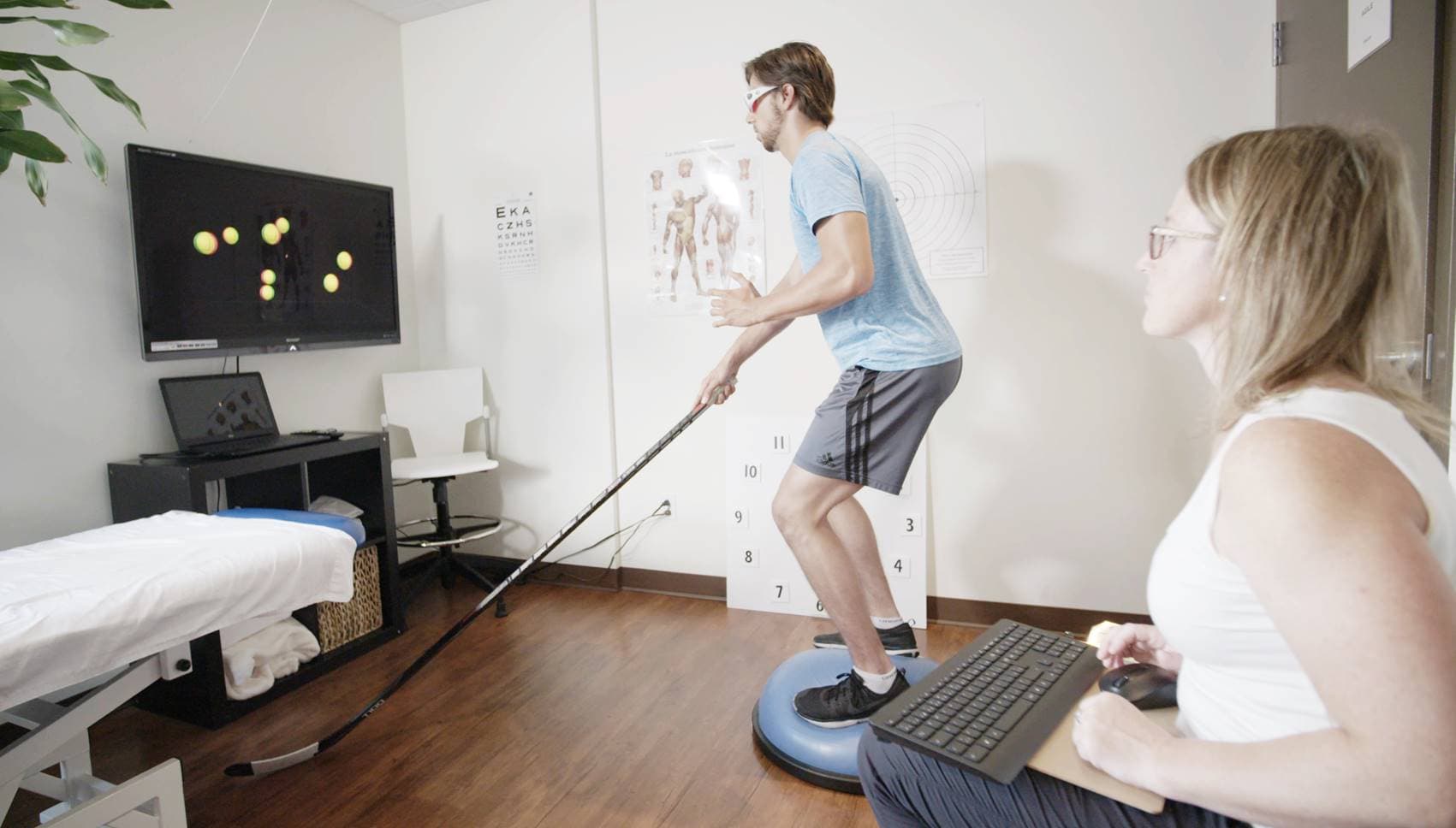 An athlete stick-handling on a balance trainer while tracking targets on a screen, with a coach running the session.